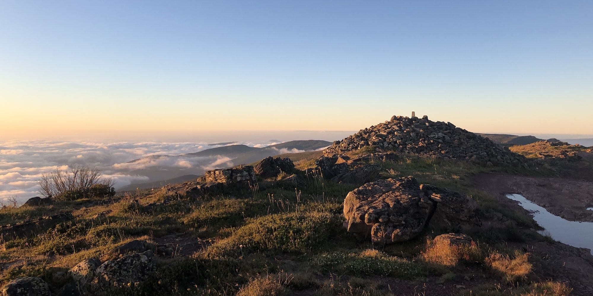 A rocky mountain-top, with other peaks poking out from above the cloudline in the background